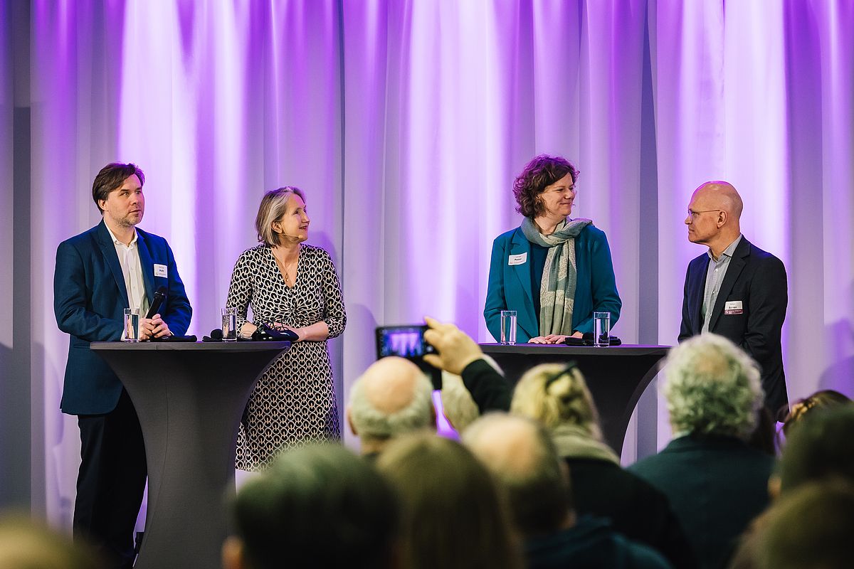 Podiumsdiskussion mit Michael Wahl, Ulrike Peters und Holger Borner. Die Teilnehmenden stehen an zwei Stehtischen. Zwischen den Stehtischen steht die Moderatorin. 